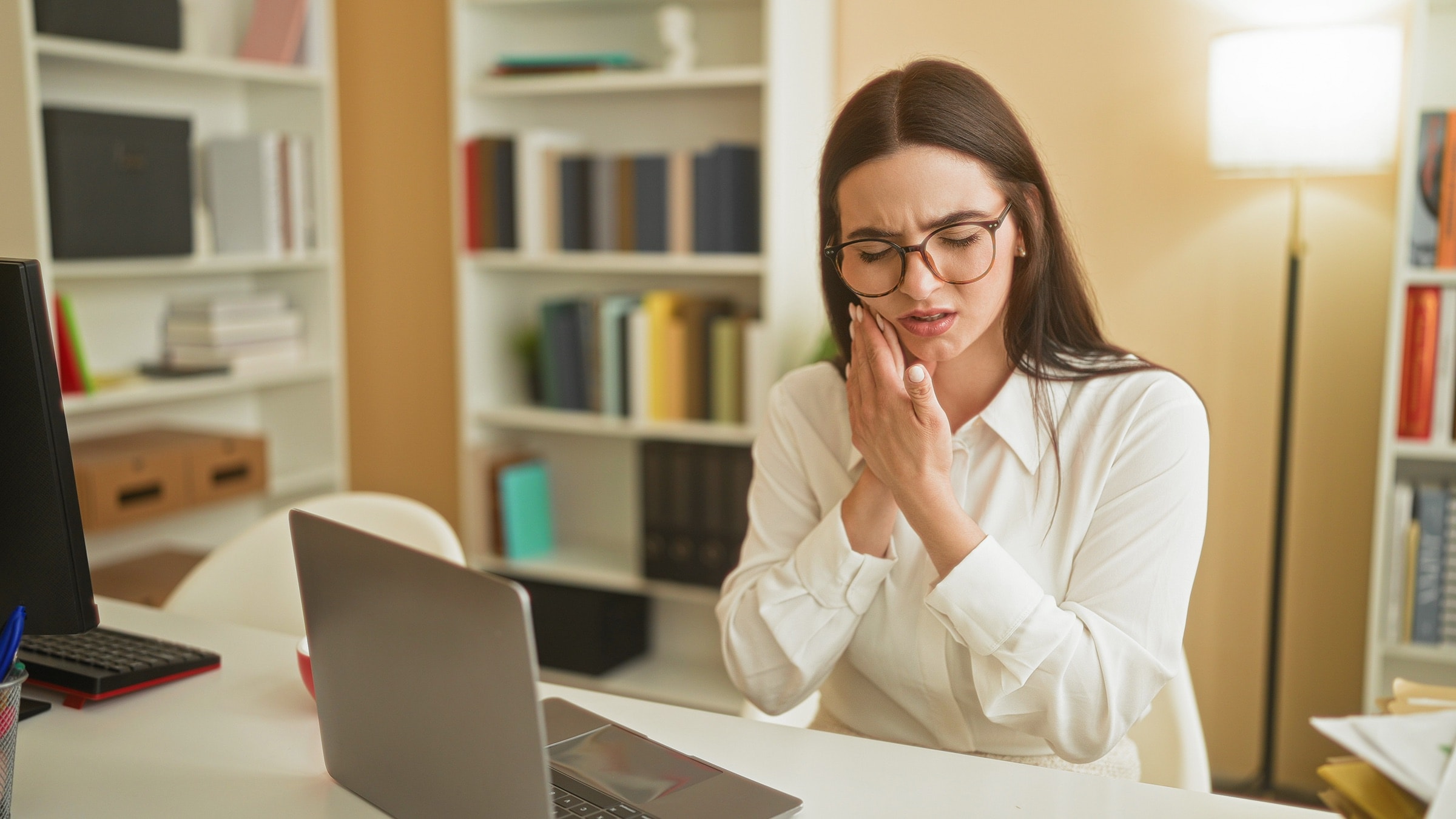 A woman at her laptop holding her jaw in pain