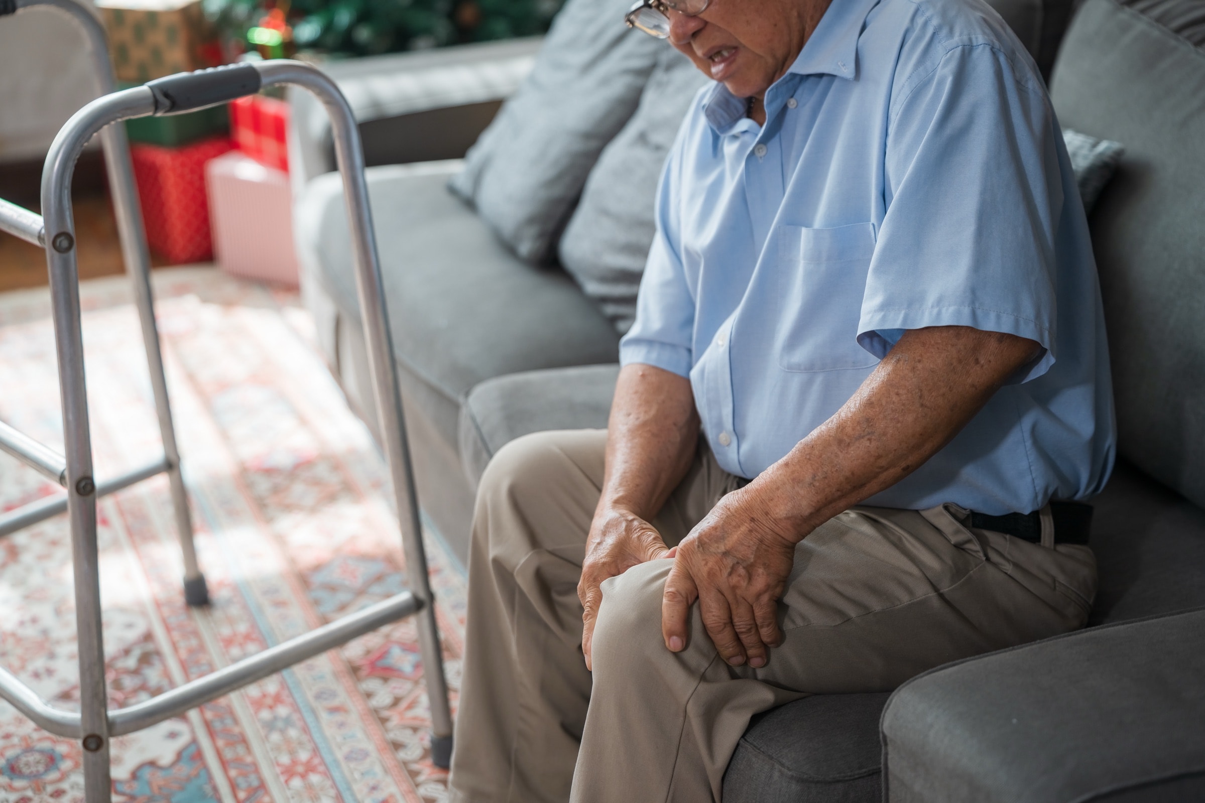 Older man on a couch holding his knee, with a walker nearby
