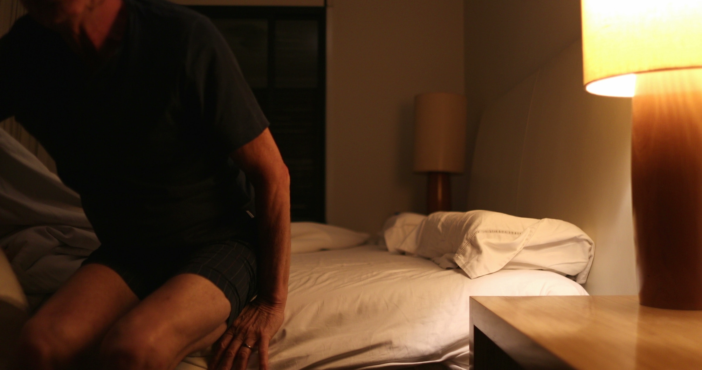 Older man sitting on the edge of his bed at night, reaching for the lamp
