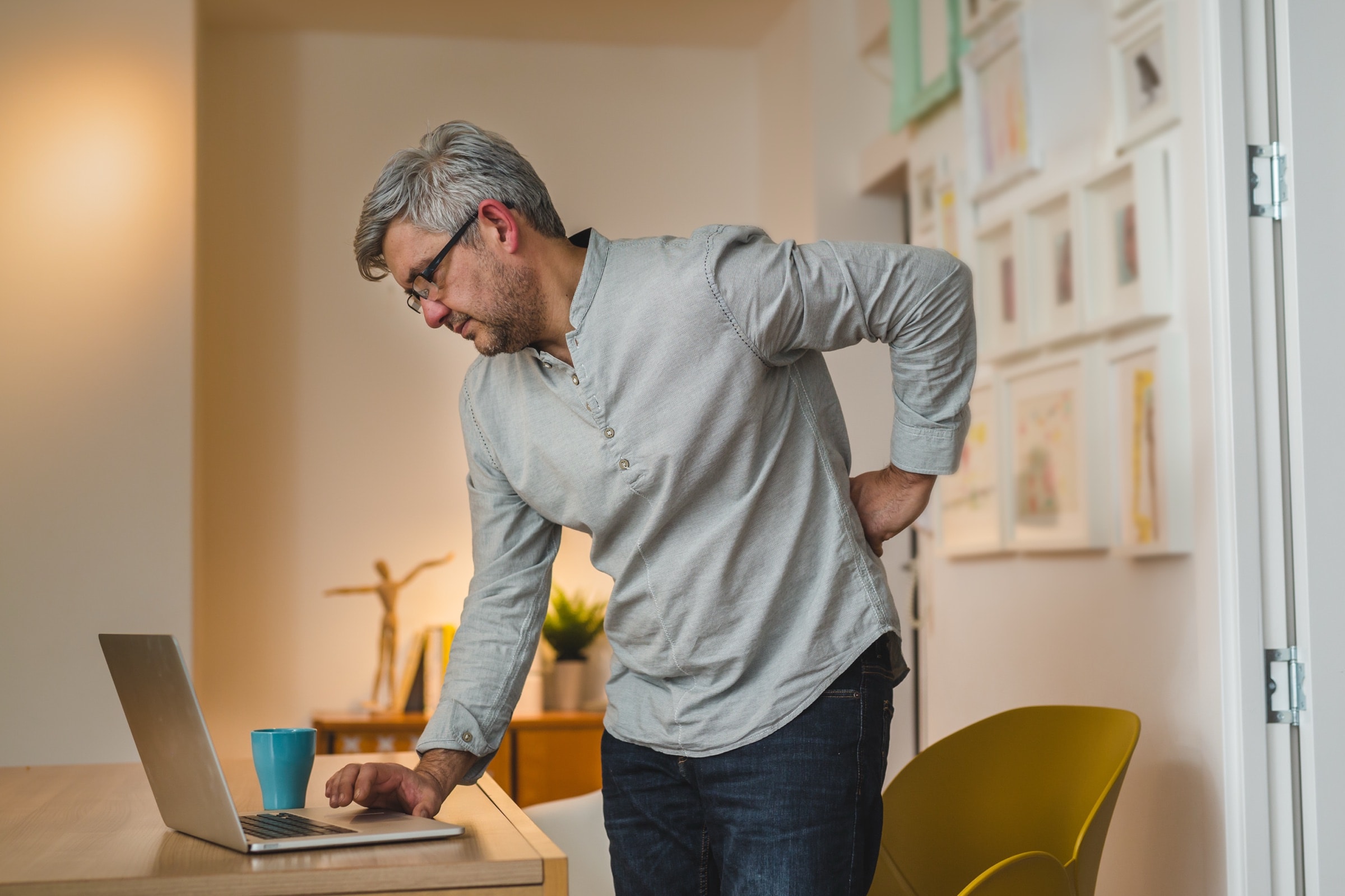 Gray-haired man getting up from his laptop, holding his lower back